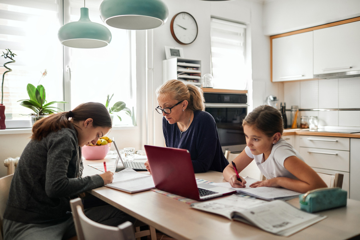 This image represents a family staying at home and working from home and doing homework in order to help curb the spread of COVID-19. The woman is reading about common investment scams that may pop up during the coronavirus pandemic.